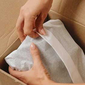 Hands wrapping a gift with white tissue paper inside a cardboard box.