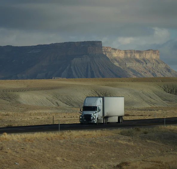 White semi-truck driving on a highway with rugged mesas and cloudy sky in the background.
