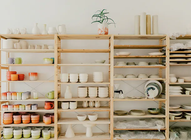 Wooden shelves displaying various handmade ceramic bowls, cups, plates, and a potted plant on top.