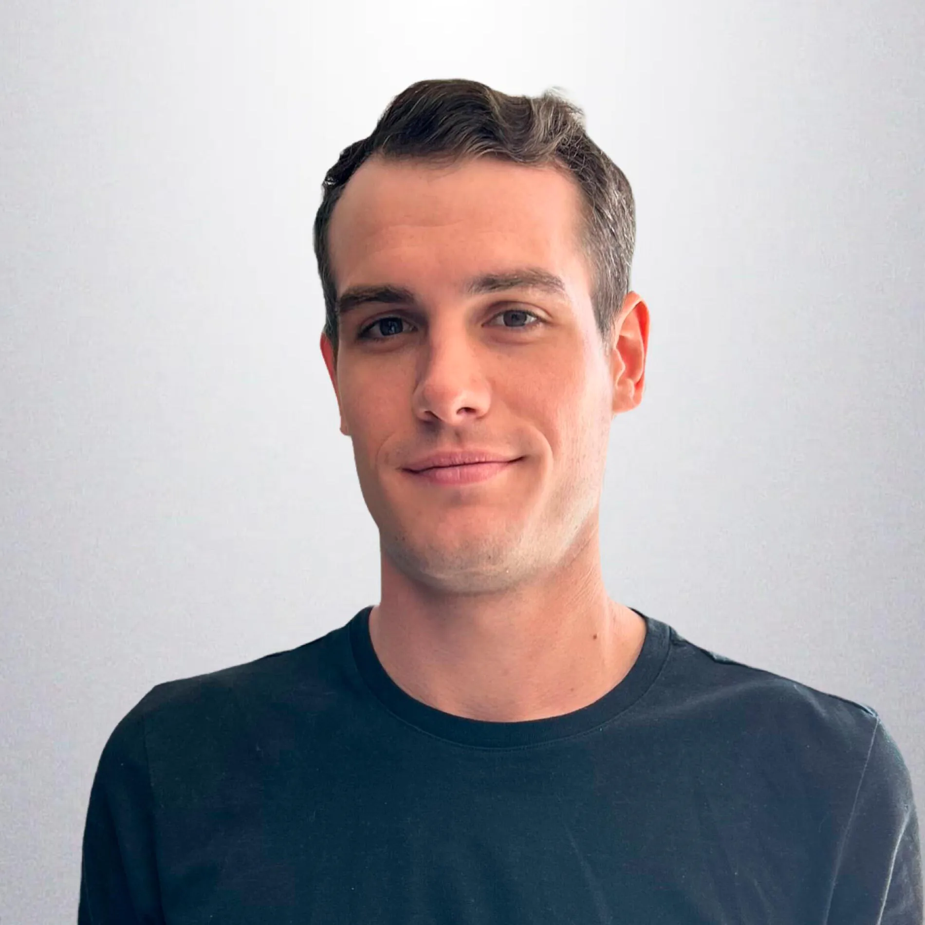 Young man with short brown hair wearing a dark crew neck shirt, smiling slightly against a plain light background.