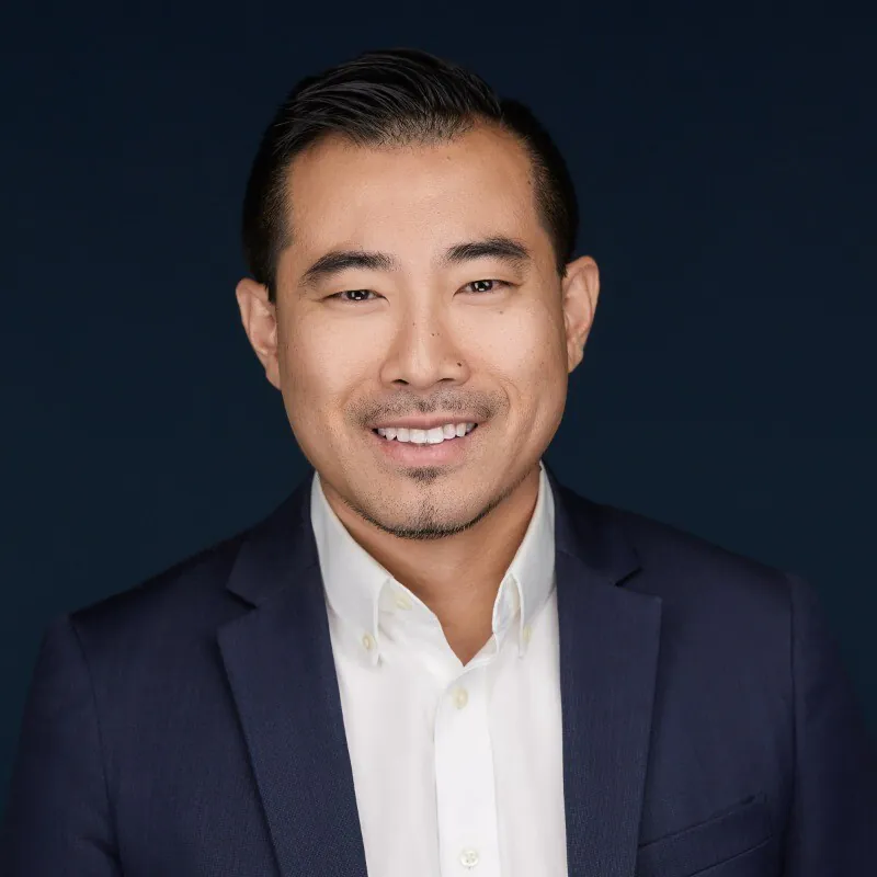 Smiling man with short black hair wearing a white shirt and dark blue blazer against a dark background.