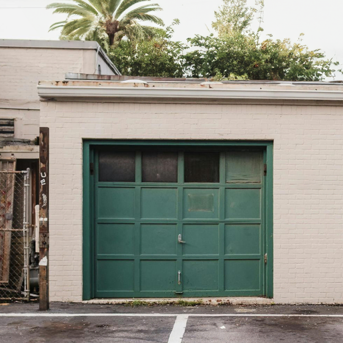 Green garage door set in a beige brick wall with a parking lot in front and palm trees visible above the roof.