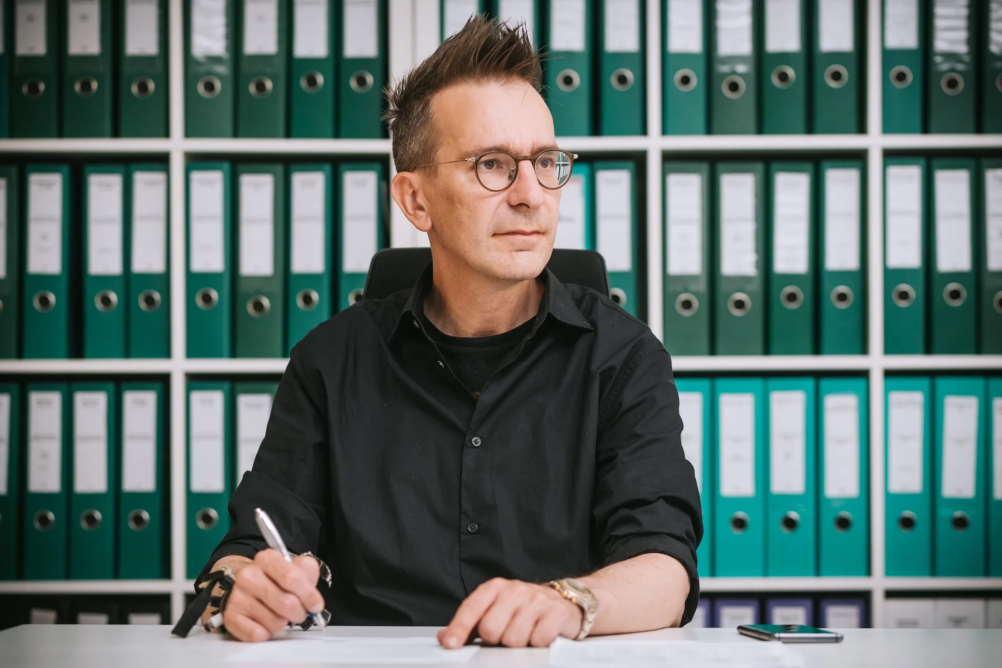 Mann mit Brille und schwarzem Hemd sitzt an einem Schreibtisch und schreibt, im Hintergrund ein Regal mit grünen Ordnern.