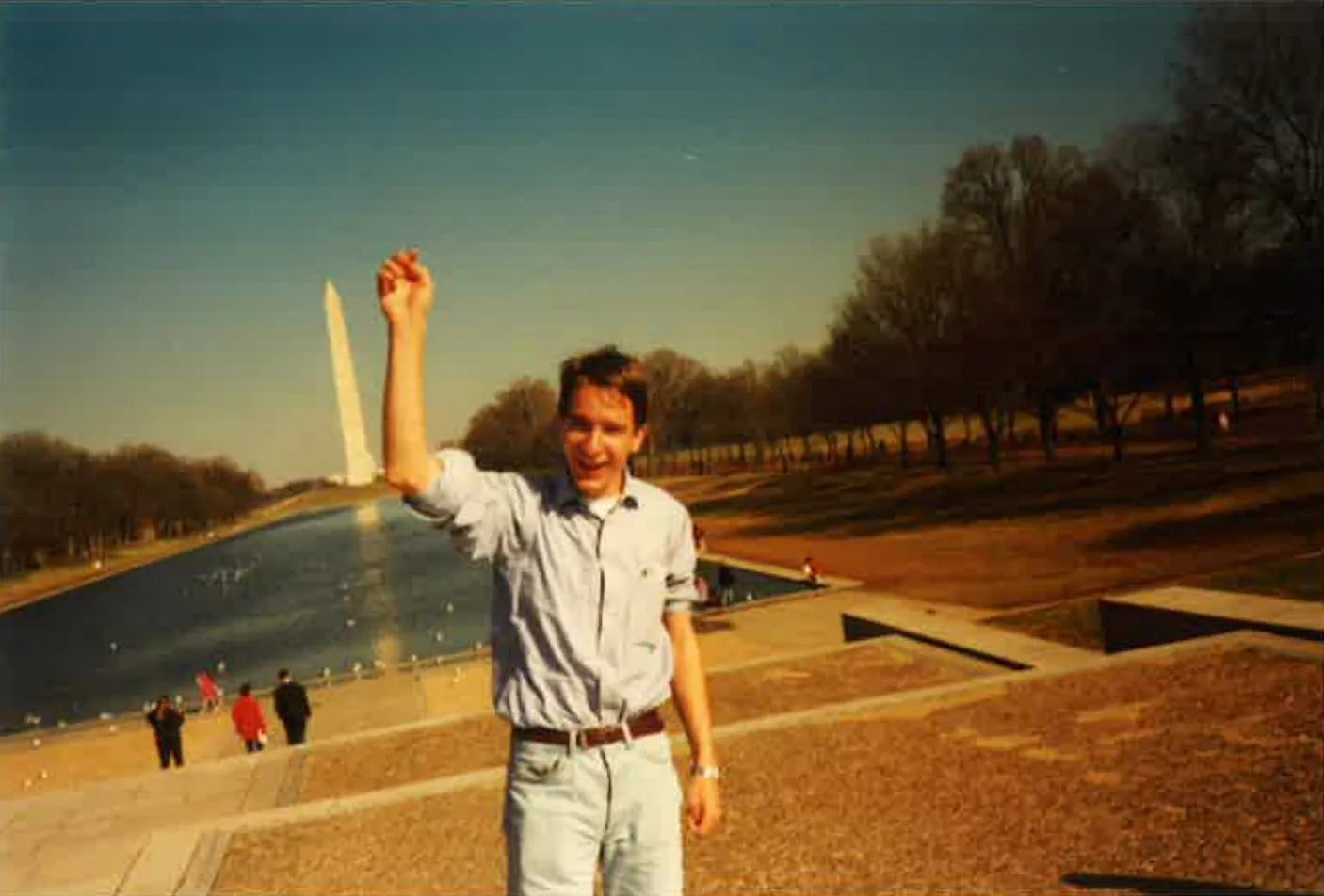 Junger Mann vor dem Washington Monument mit gehobener rechter Hand und lächelndem Gesichtsausdruck am Reflecting Pool.