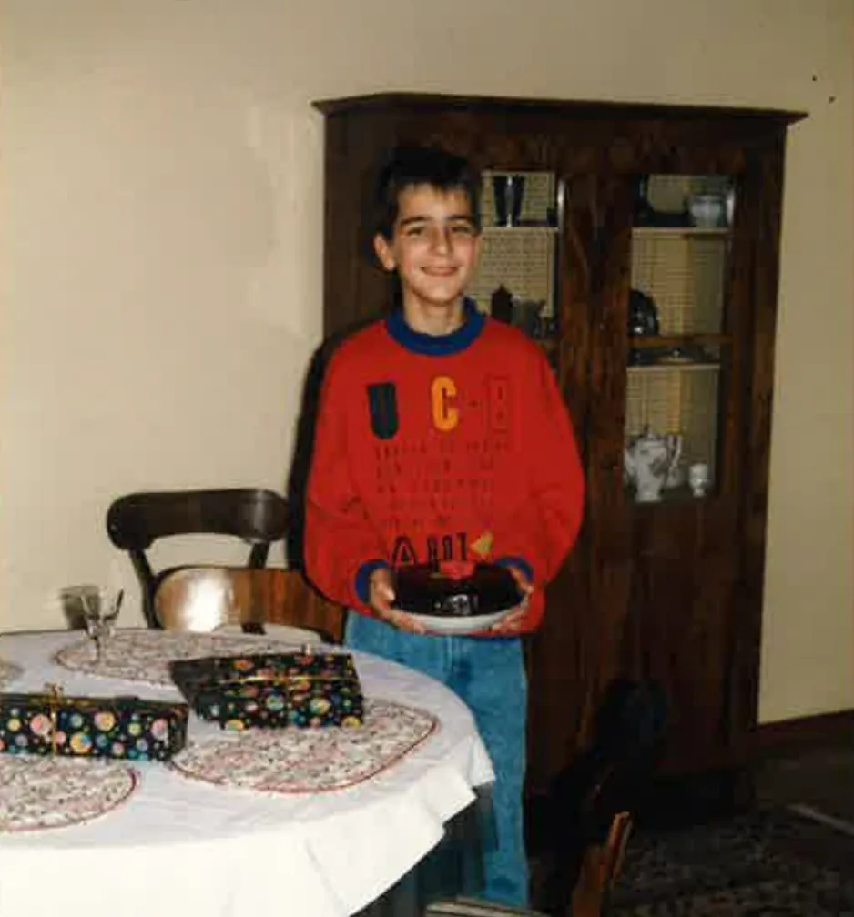 Lächelnder Junge in rotem Pullover und blauen Jeans hält einen kleinen Schokoladenkuchen in einem Teller in einem Esszimmer.