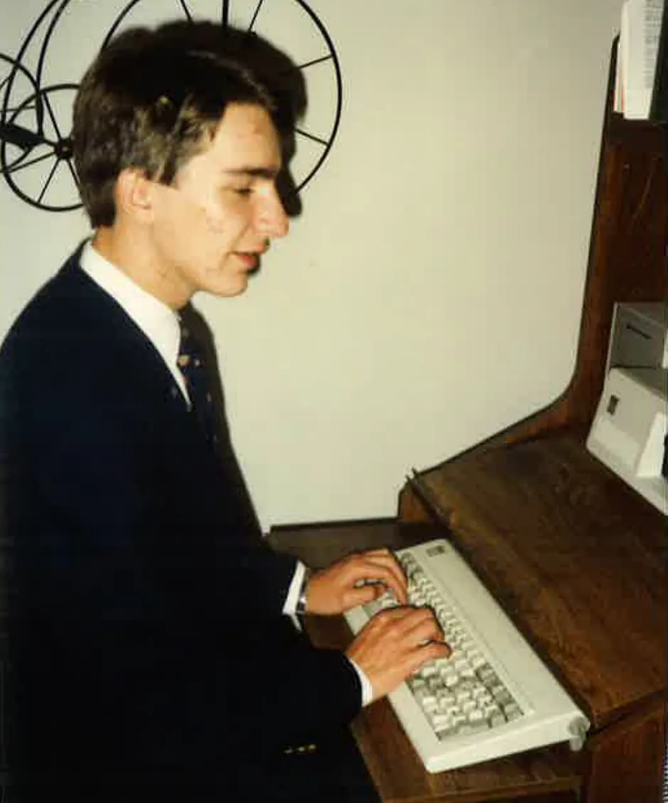 Junger Mann in dunklem Anzug tippt an einer Tastatur eines alten Computers auf einem Holzschreibtisch.