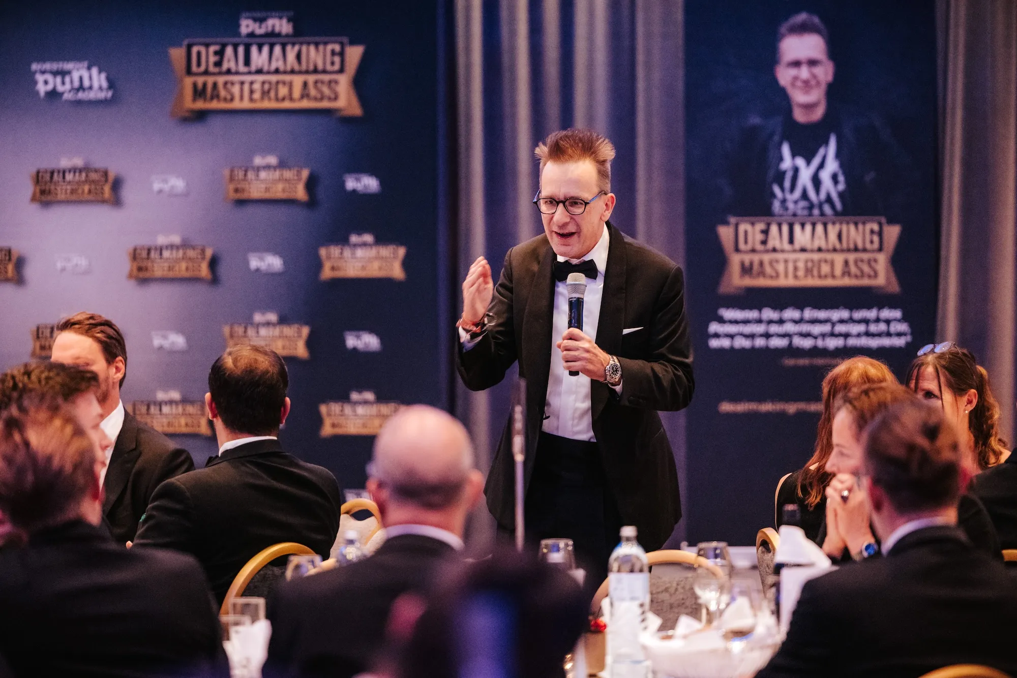 Mann im schwarzen Anzug und Fliege hält eine Rede mit Mikrofon bei der Dealmaking Masterclass vor Publikum.