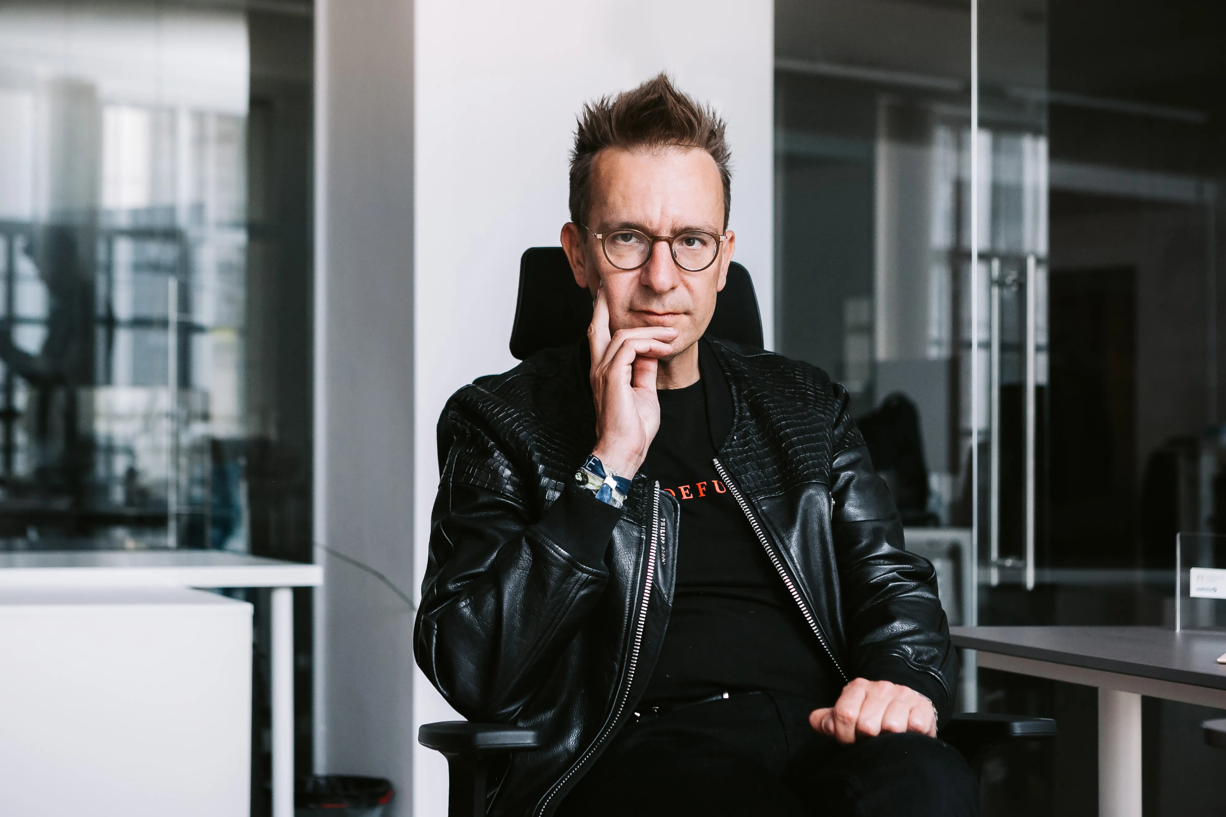 Mann mit Brille und schwarzer Lederjacke sitzt nachdenklich auf einem Bürostuhl in einem modernen Büro.