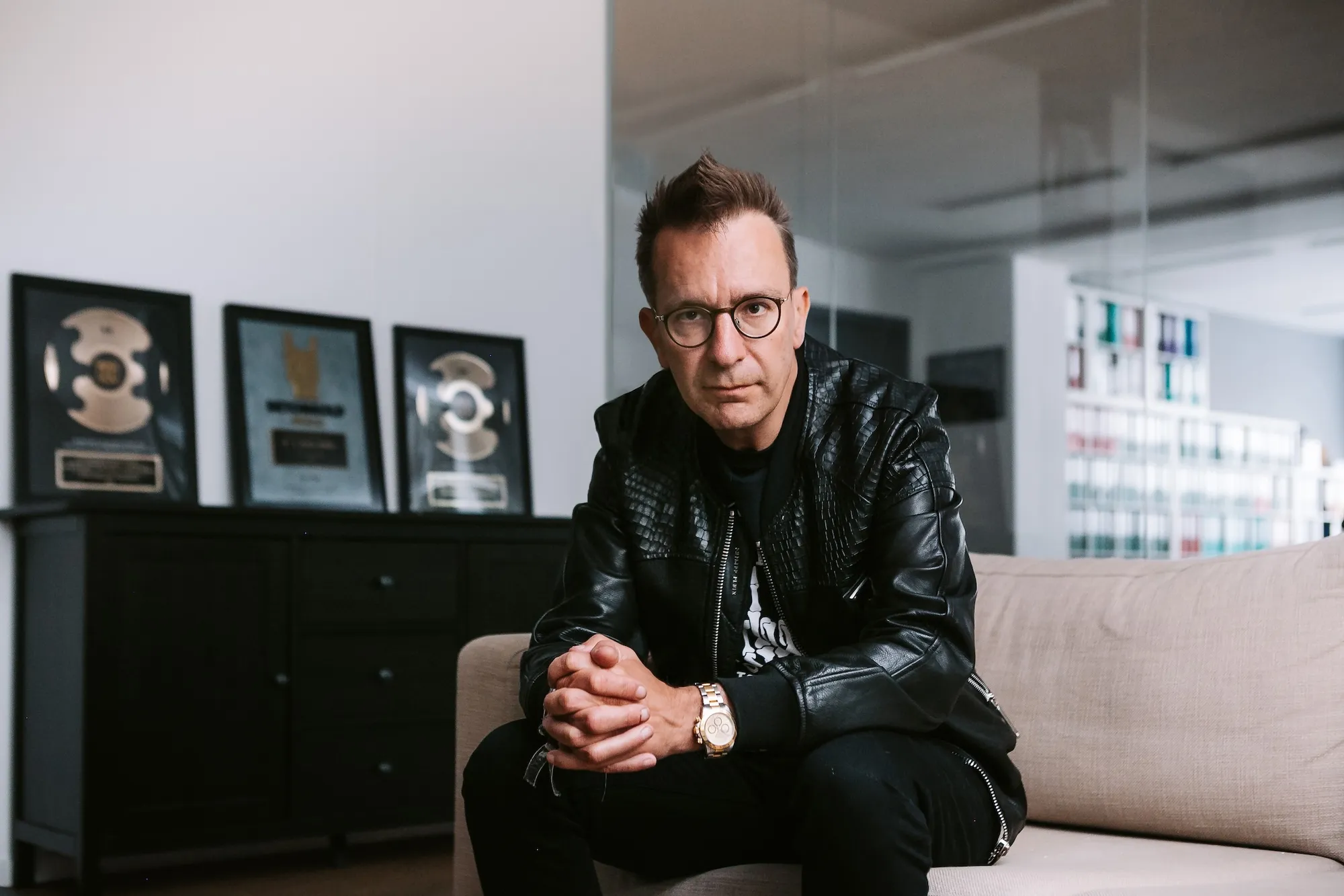 Mann mit Brille und schwarzer Lederjacke sitzt auf einem Sofa in einem modernen Büro mit Musikplaketten im Hintergrund.