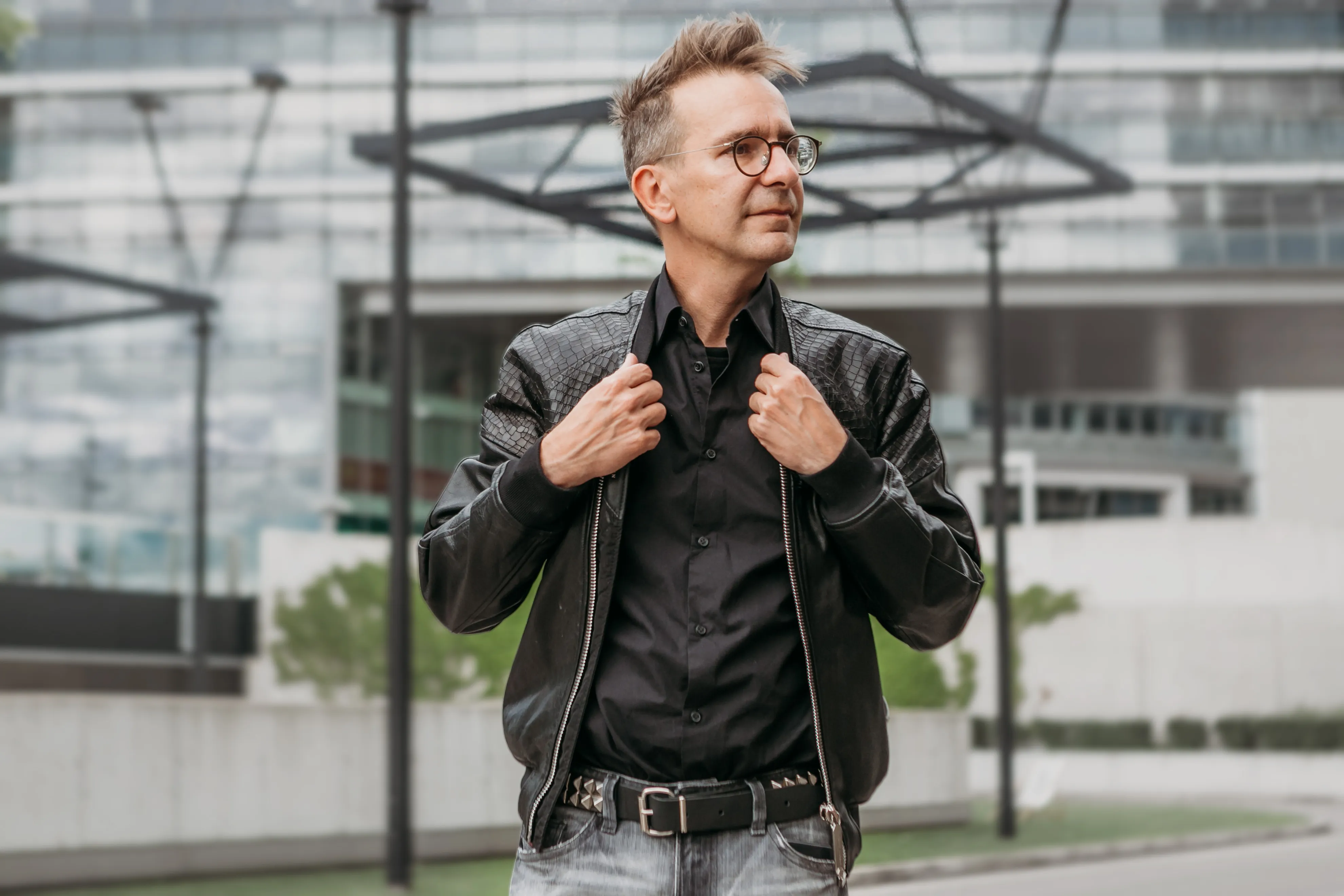 Mann mit Kurzhaarschnitt und Brille trägt schwarze Lederjacke und Hemd vor modernem Bürogebäude.