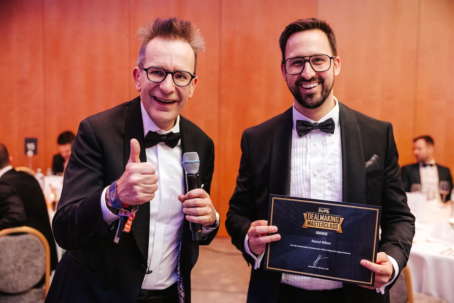 Zwei Männer in Smoking, einer hält ein Mikrofon und zeigt Daumen hoch, der andere hält eine Urkunde für die Teilnahme an einer Dealmaking Masterclass.