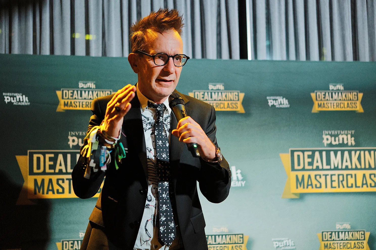 Mann mit Brille und Krawatte mit Totenkopf-Motiv hält Mikrofon und spricht bei einem Dealmaking Masterclass Event.