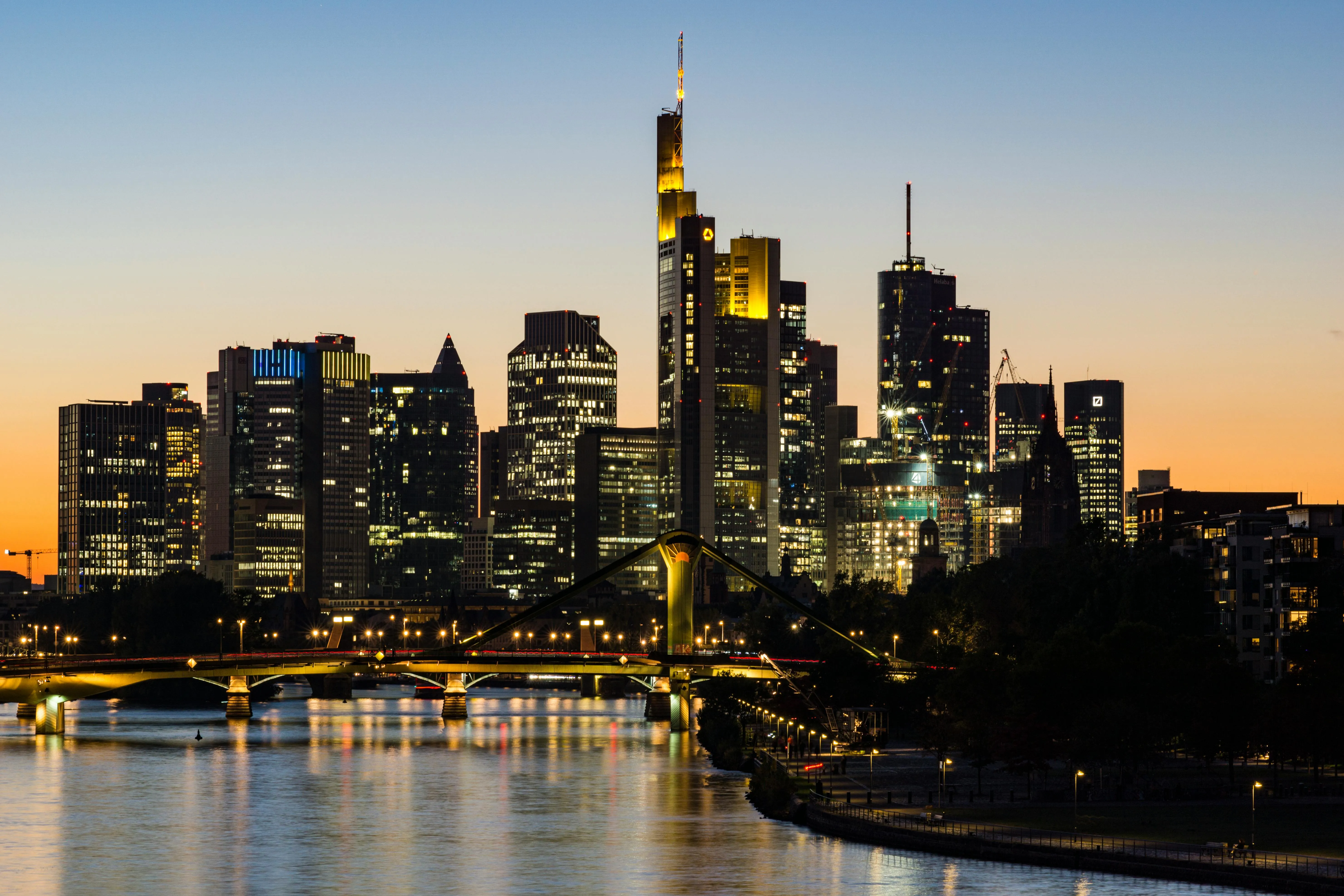 Skyline von Frankfurt am Main bei Sonnenuntergang mit erleuchteten Wolkenkratzern und Fluss im Vordergrund.