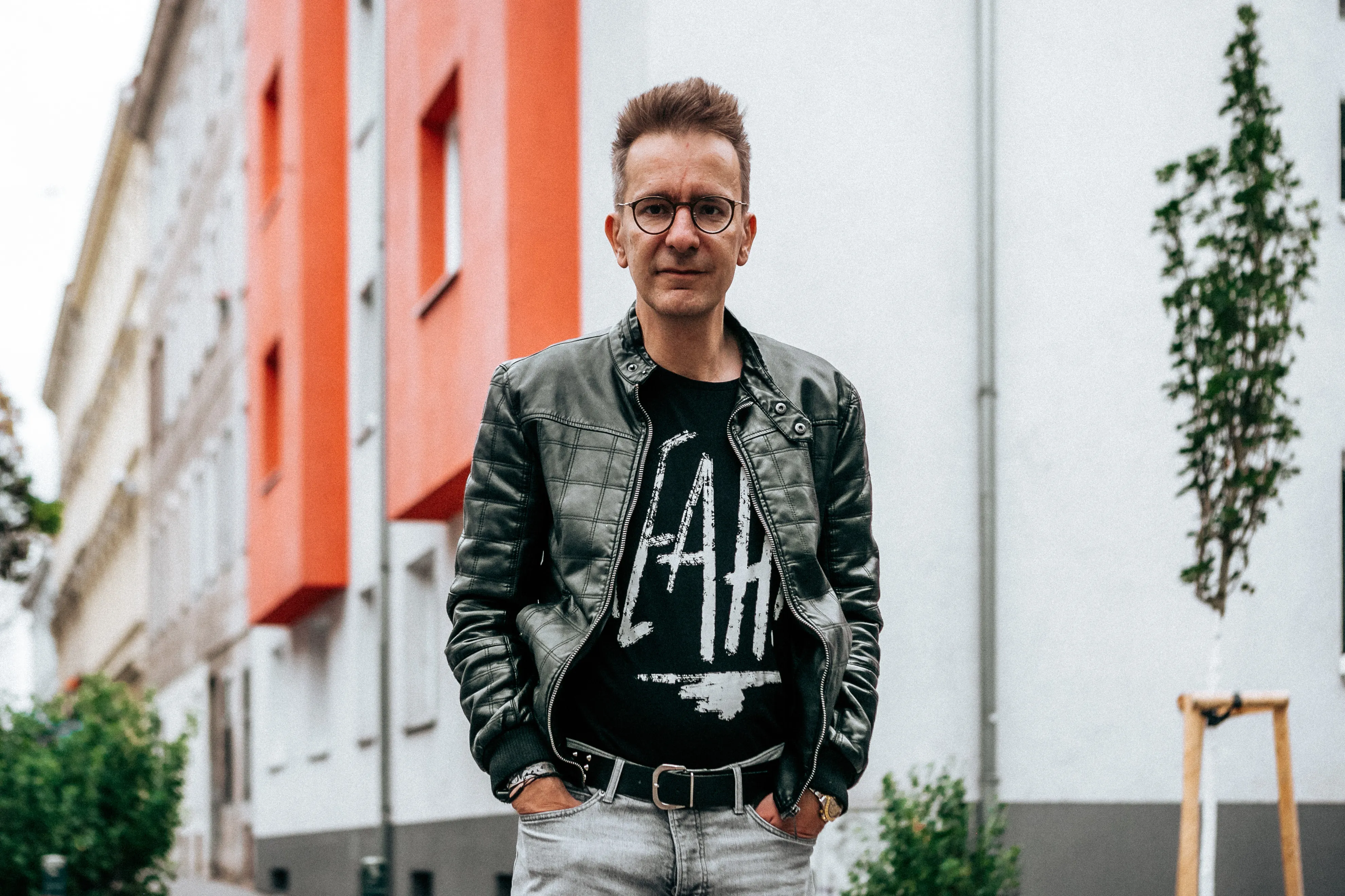 Gerald Hörhan mit Brille, schwarzer Lederjacke und T-Shirt vor einem Gebäude mit roten und weißen Wänden.
