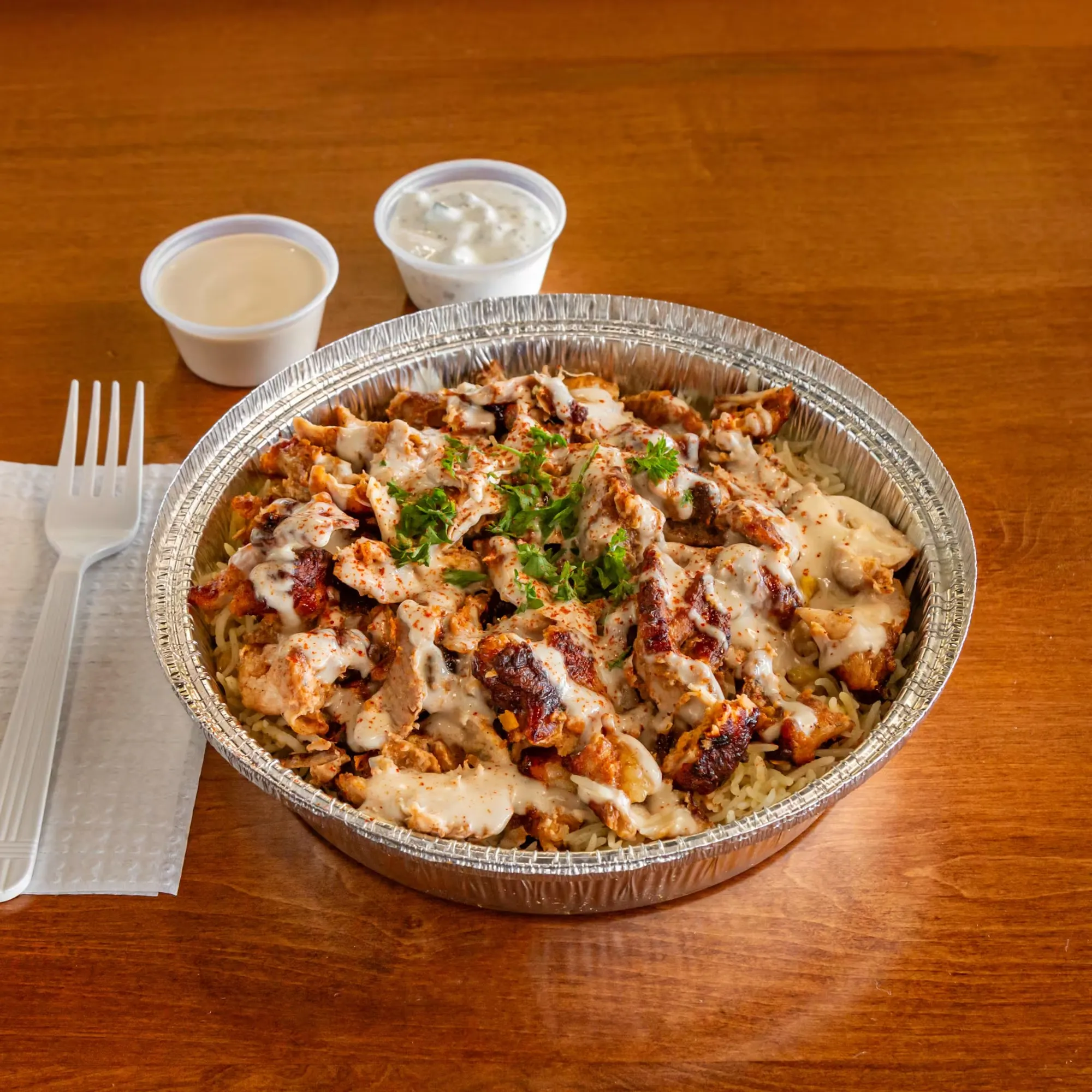 A foil container of chicken with a side of sauce.