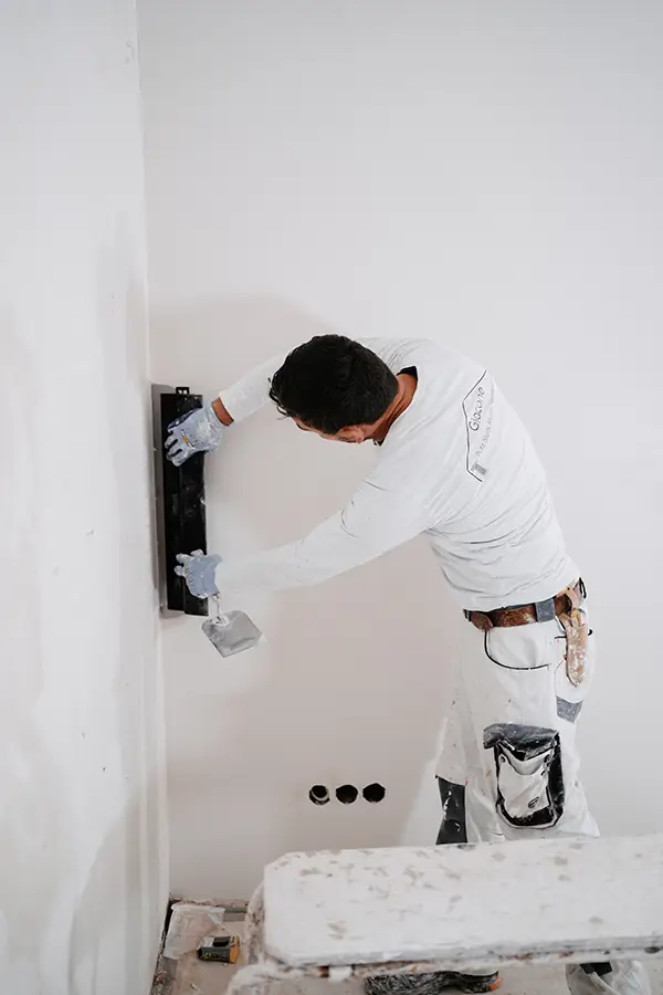 Worker in white clothing smoothing a drywall wall with a trowel in an interior room.