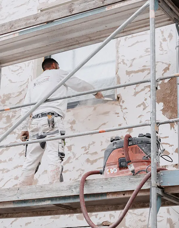 Painter on scaffolding applying white paint to a stucco wall near a window.
