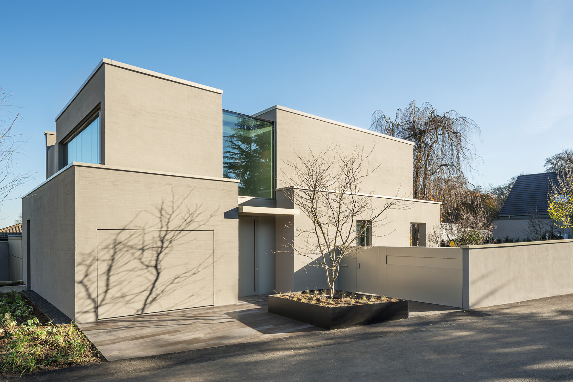 Modern beige house with flat roof, large windows, planter with leafless tree, and shadow on garage door.