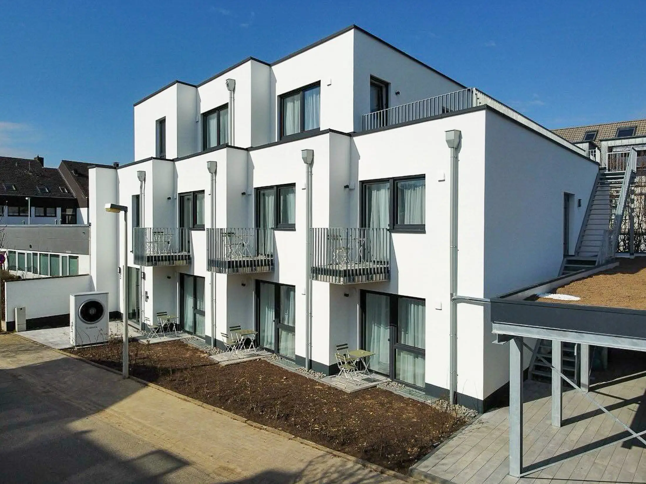Modern white building with balconies, small patio tables, and clear blue sky background.