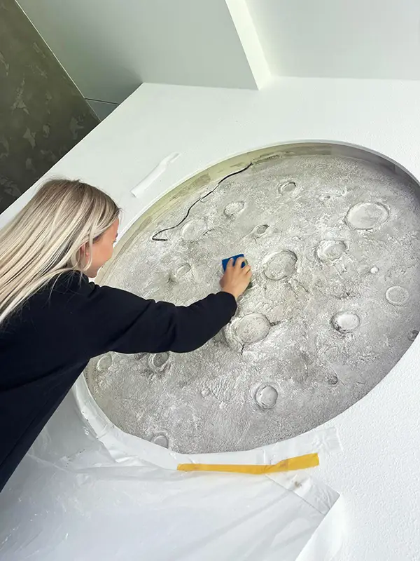 Person cleaning or working on a round textured wall art piece with craters resembling the moon.