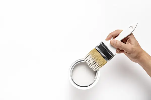 Hand holding a white paintbrush gently dipped in a small open can of white paint on a white surface.