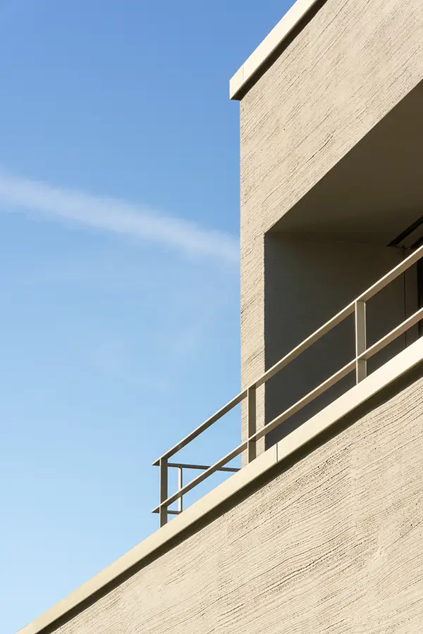 Modern beige building corner with metal railing against a clear blue sky.