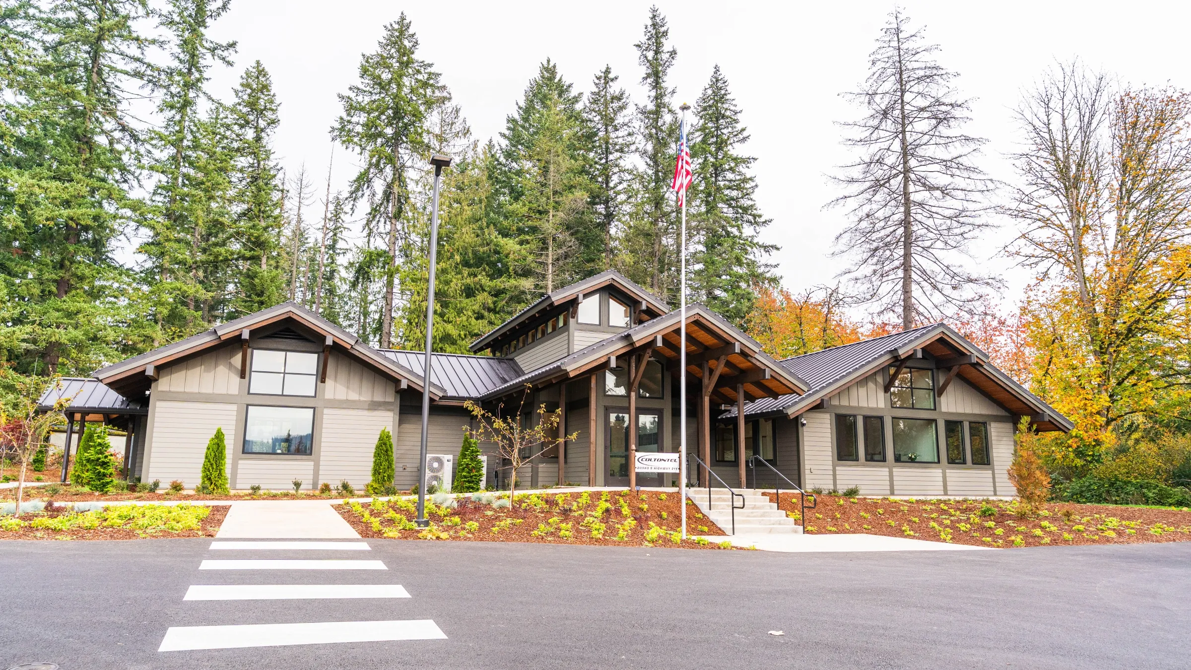 Modern single-story building with peaked roofs, large windows, and an American flag pole in front, surrounded by trees.