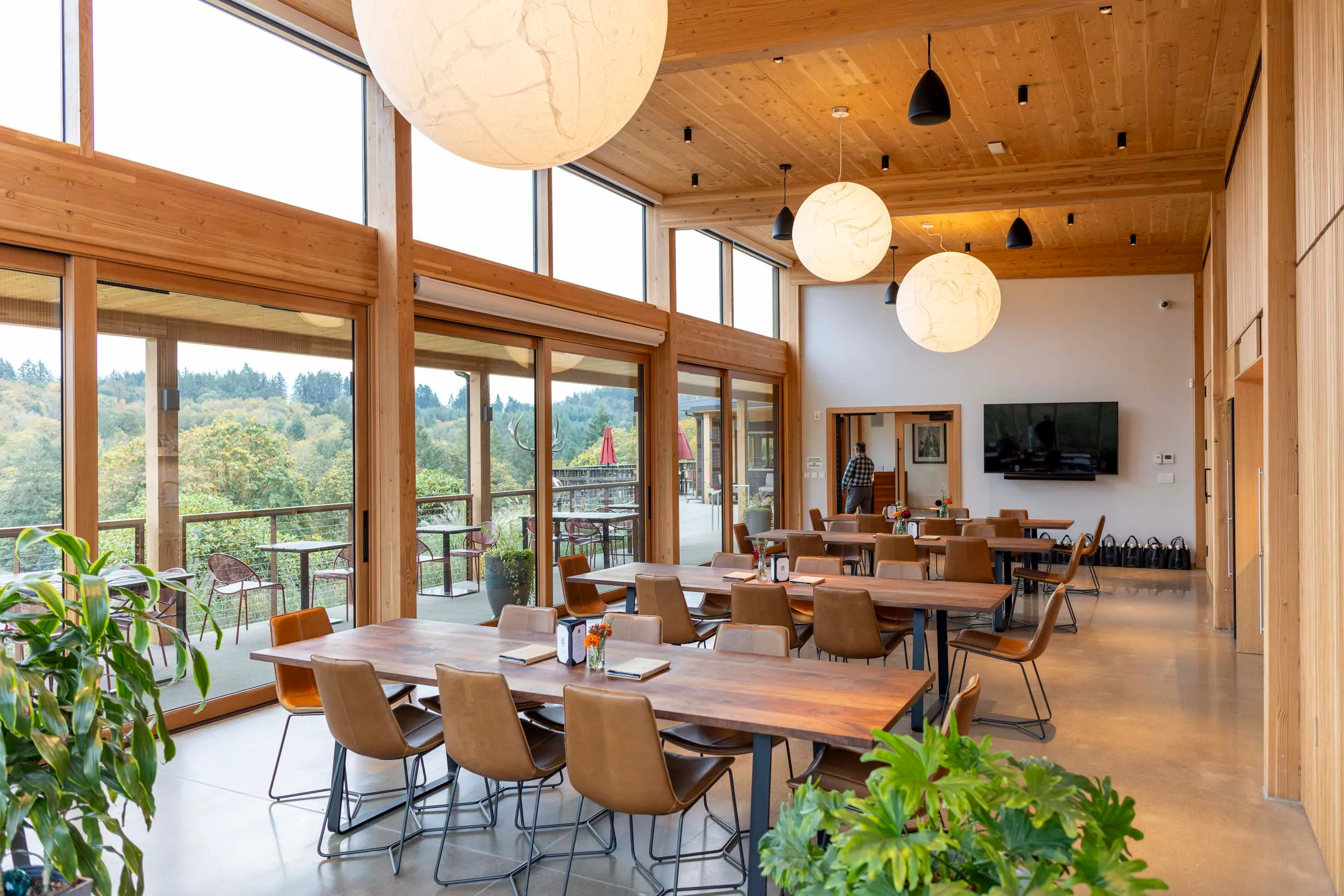 Spacious dining area with wooden tables and chairs, large windows overlooking a forested landscape, and hanging spherical light fixtures.