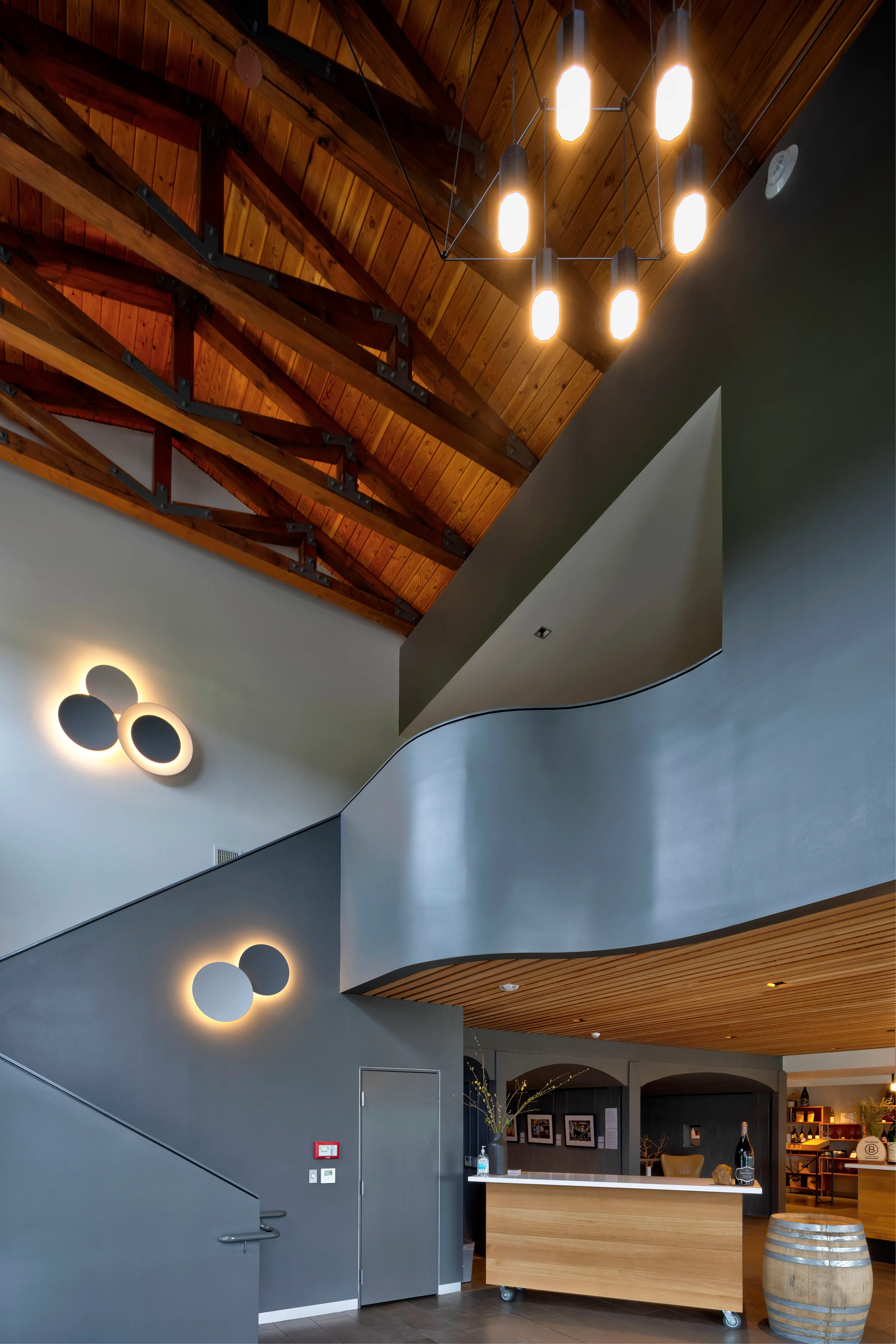 Modern interior with curved gray staircase, wooden ceiling with exposed beams, lit wall sconces, a wooden reception desk, and wine barrel.