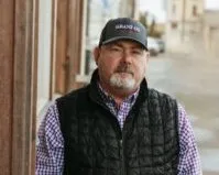 Man with a gray beard wearing a black cap, checkered shirt, and black vest standing outdoors near a wooden wall.