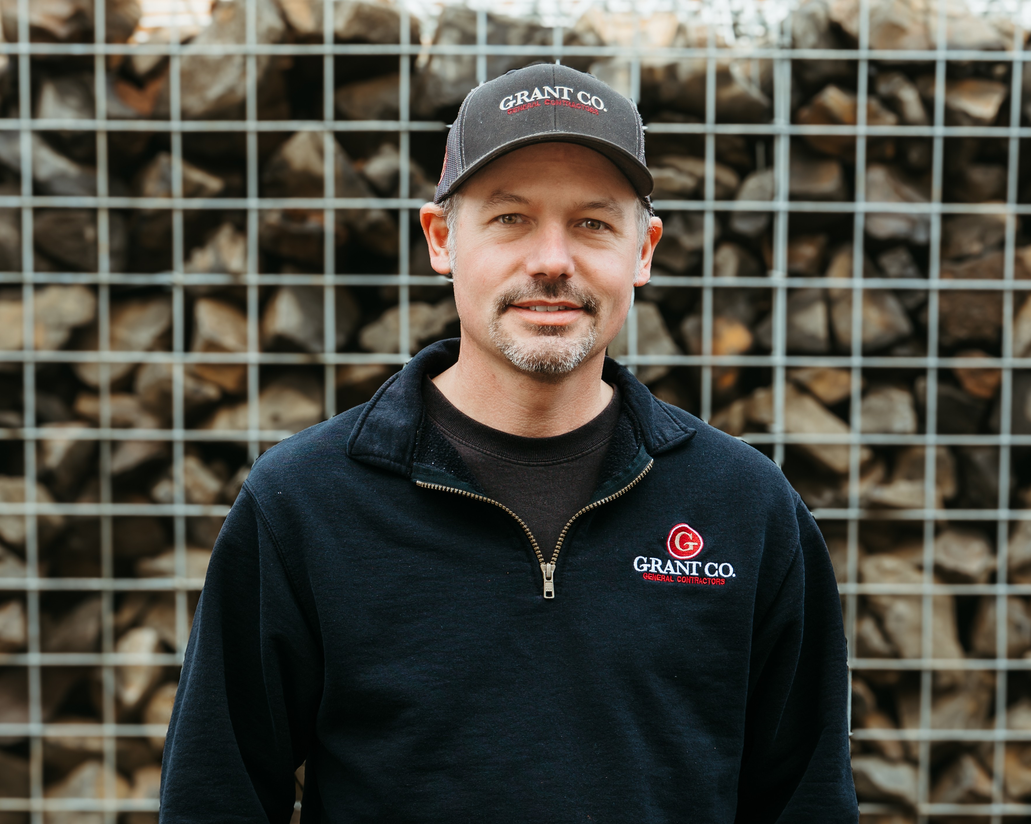 Man wearing a Darkstone cap and a black zip-up jacket standing in front of stacked firewood behind a wire fence.