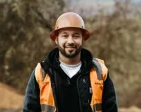 Man wearing an orange safety helmet and reflective vest smiling outdoors with blurred trees in the background.