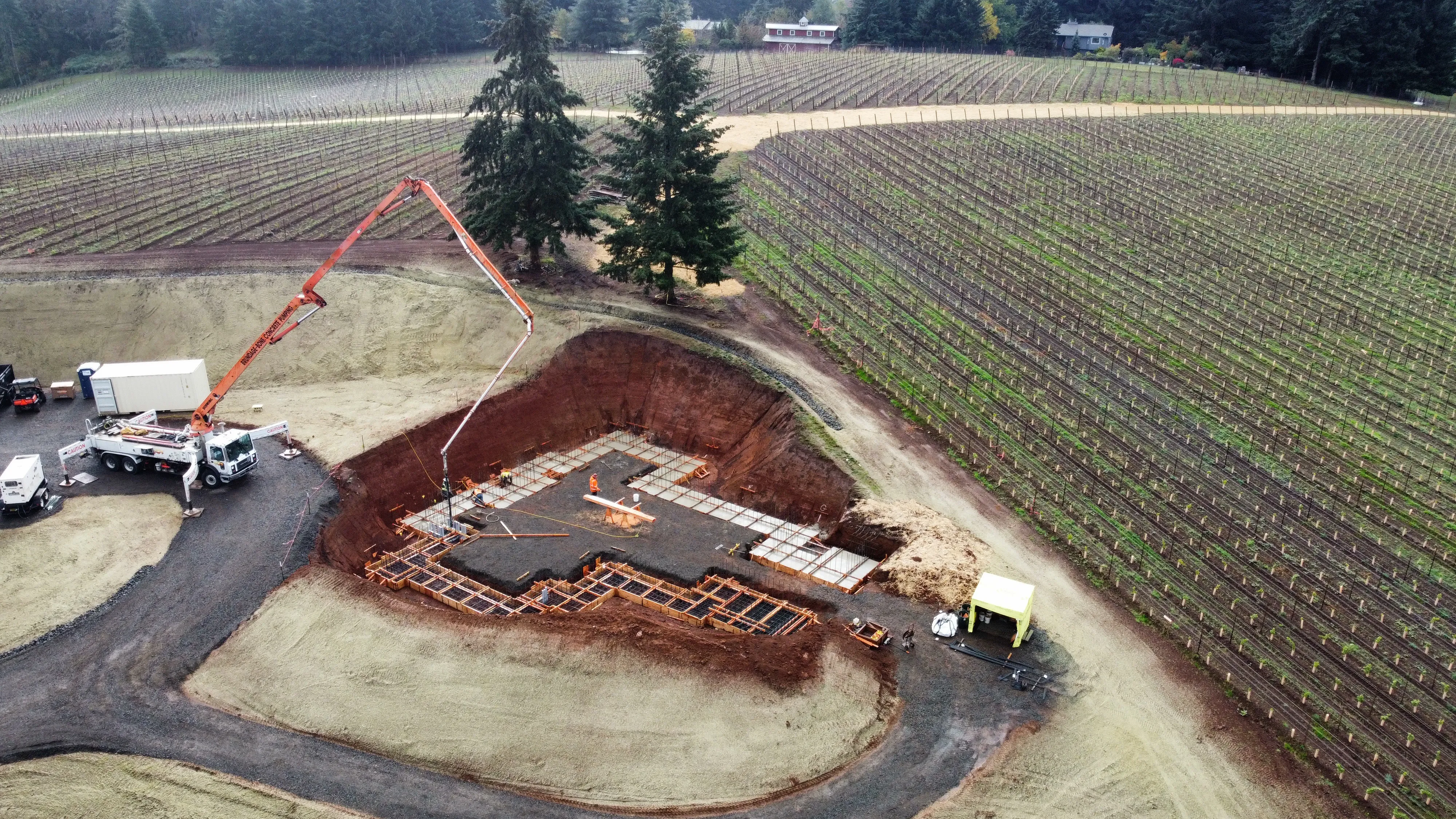 Aerial view of a construction site with foundation work in progress near a vineyard with rows of grapevines and surrounding trees.
