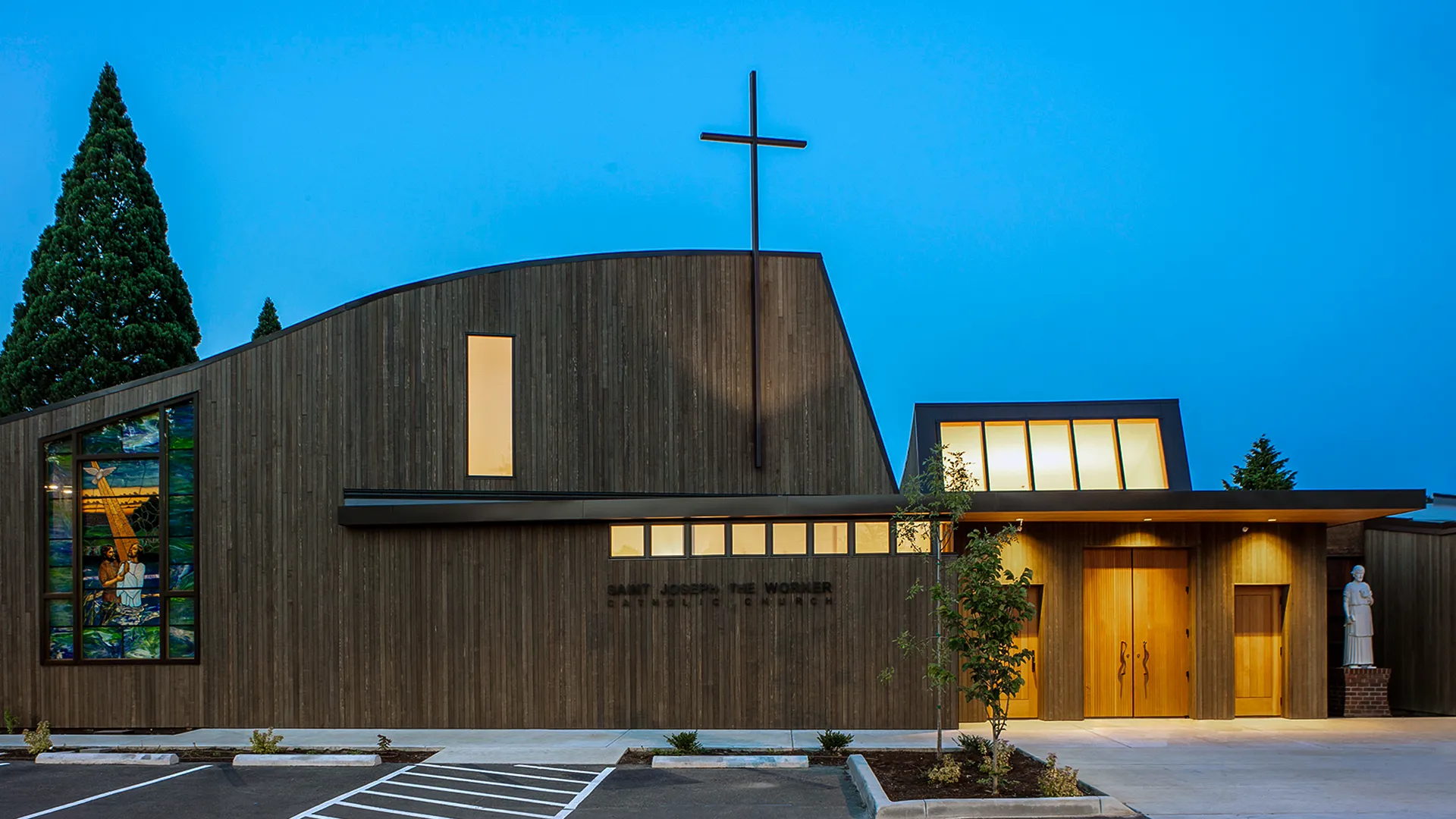 Modern church building with a tall cross, stained glass window, wooden doors, and a statue outside during twilight.