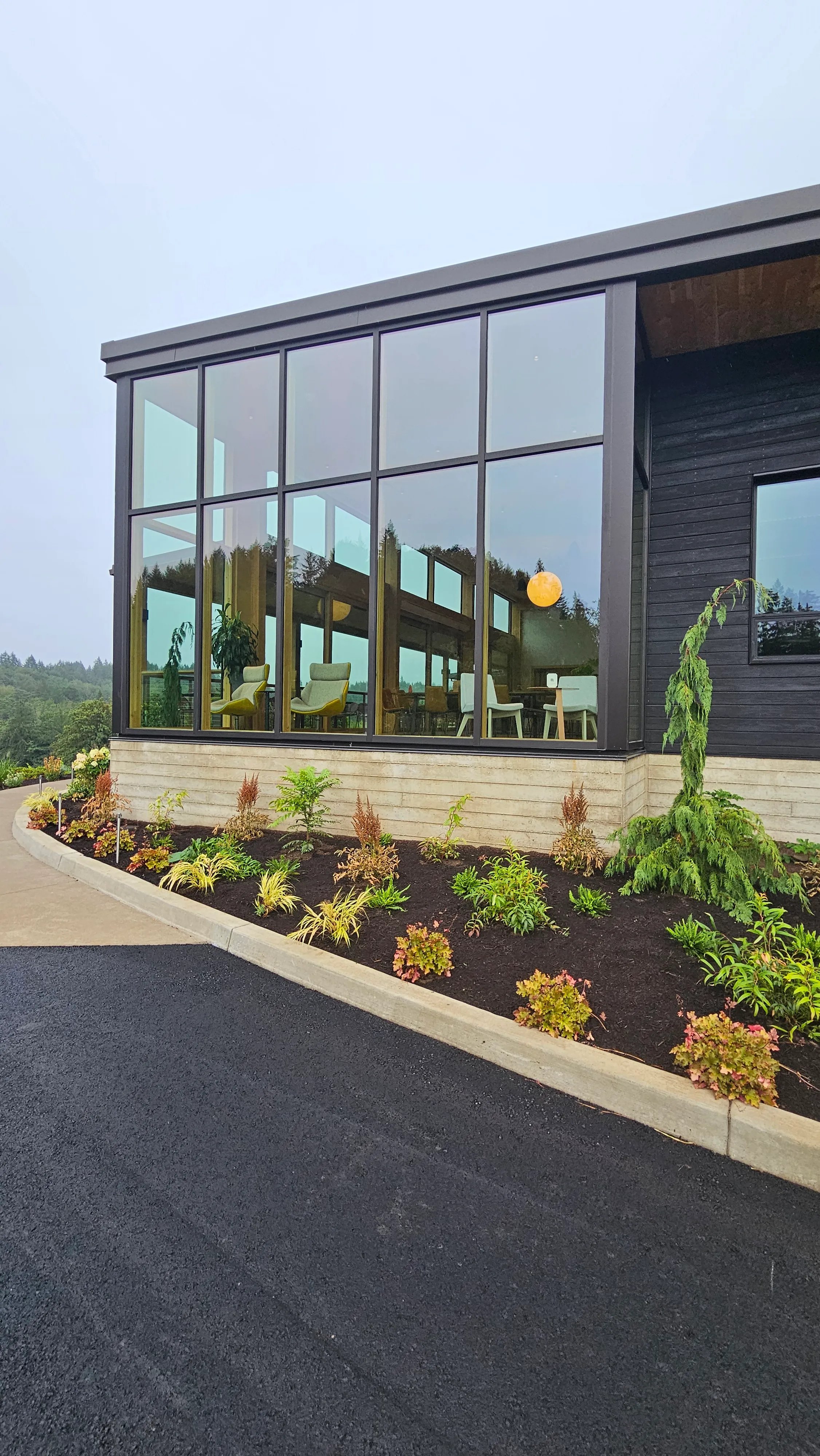 Modern building corner with large floor-to-ceiling windows showing indoor chairs and a round hanging light, surrounded by landscaped plants and a paved driveway.