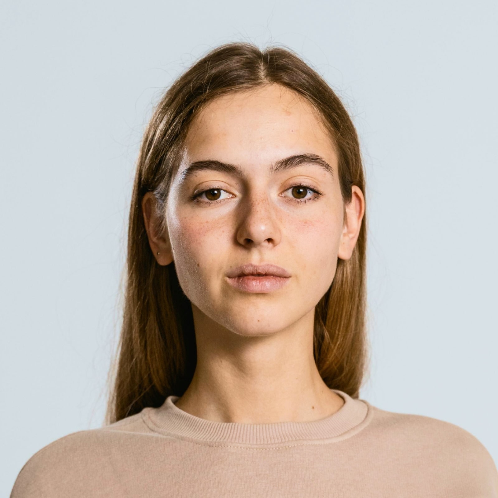 Young woman with long brown hair wearing a beige top, looking directly at the camera against a light background.