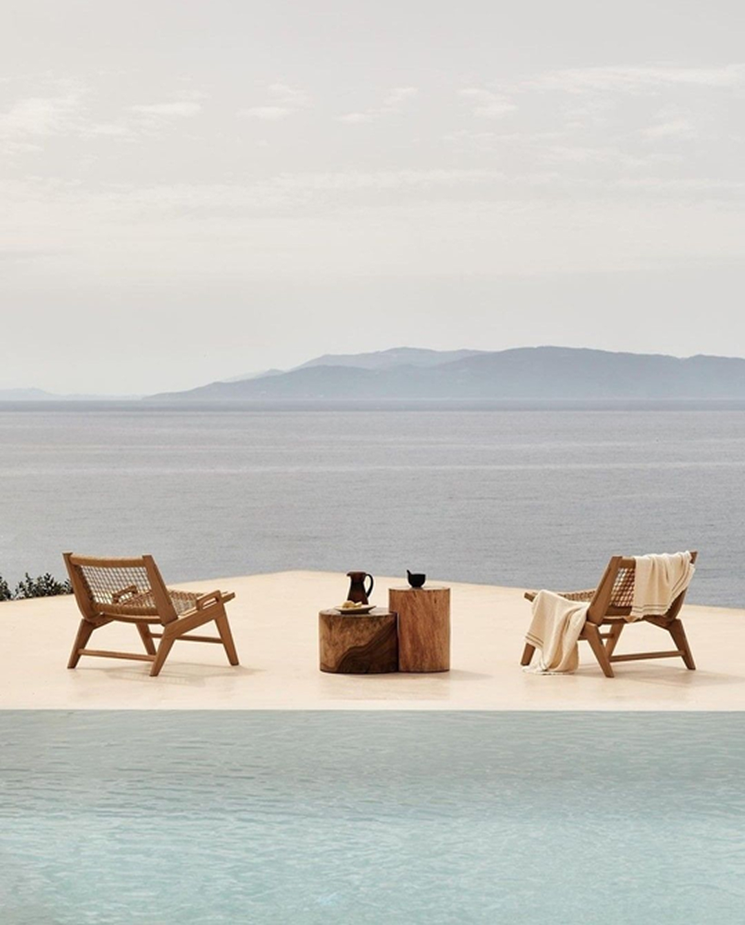 Two wooden lounge chairs with blankets beside a pool overlooking a serene sea and distant mountains.