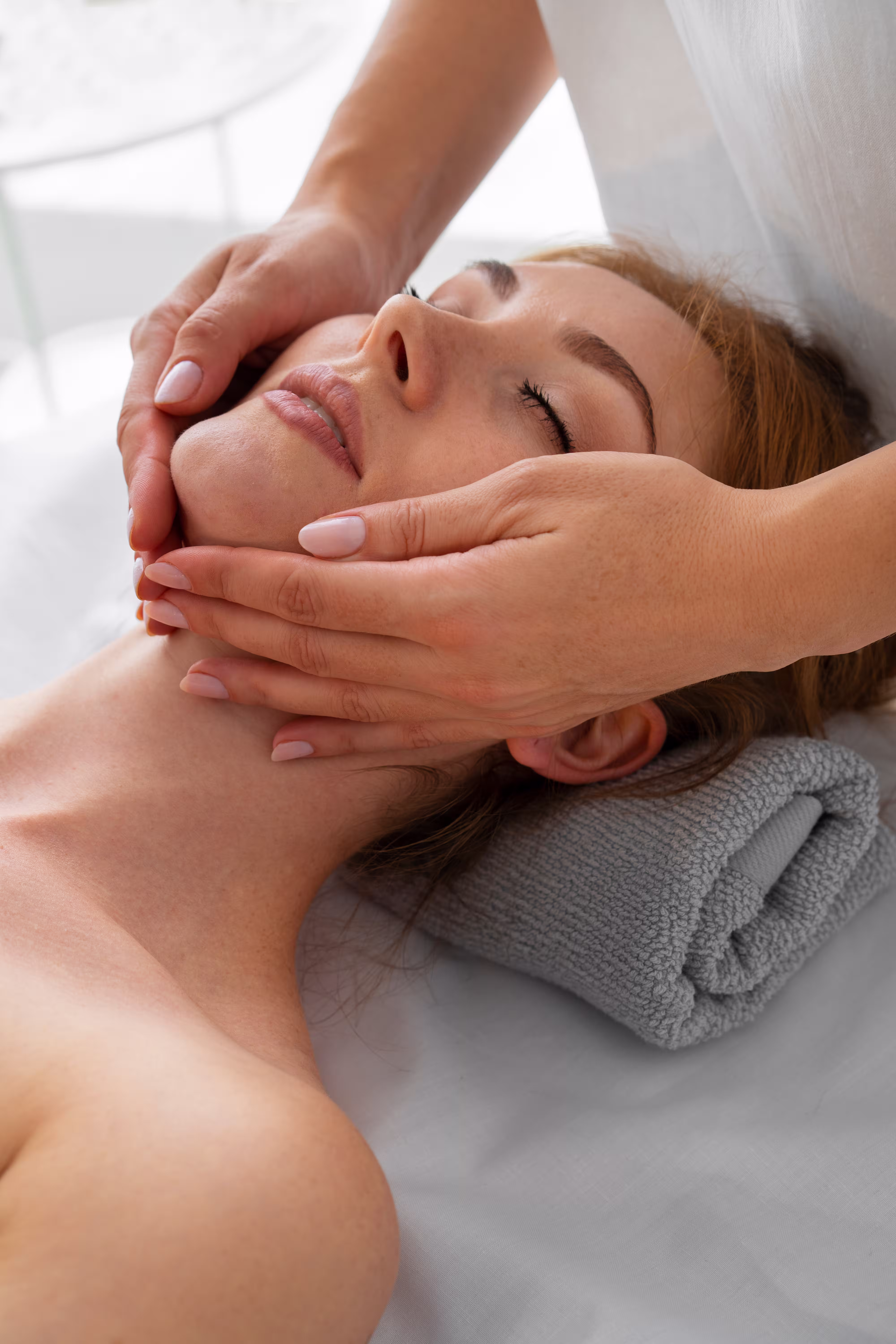 Relaxed woman receiving a gentle facial massage while resting her head on a rolled grey towel.