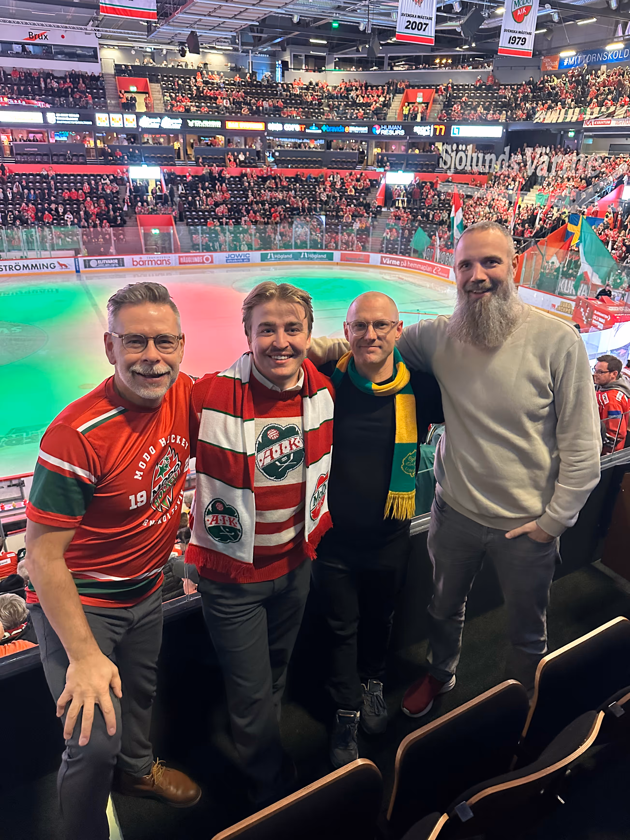 Four men posing and smiling inside a crowded ice hockey arena, with some wearing sports scarves and jerseys.