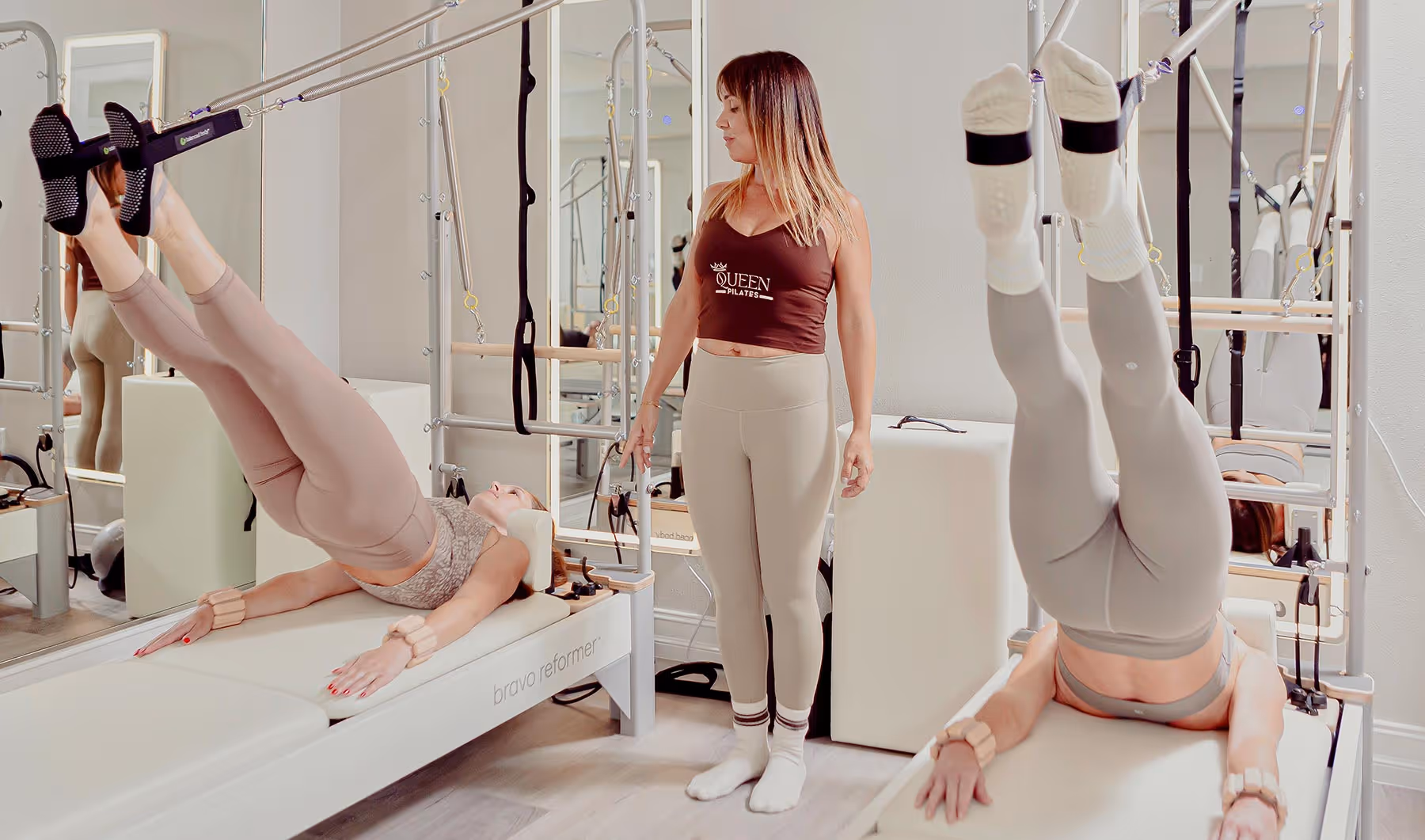 Two women perform leg exercises on Pilates reformer machines while an instructor watches in a studio.