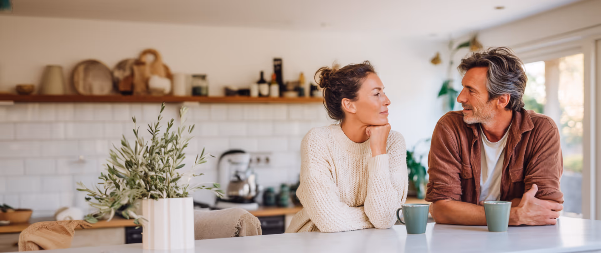 Man and woman sitting at a kitchen counter, smiling and looking at each other with coffee mugs in front of them.