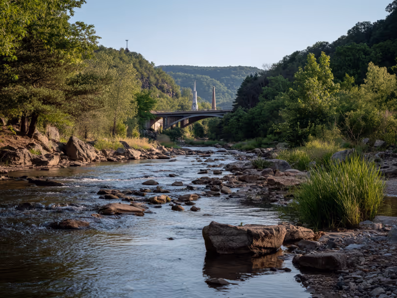Shallow river with rocks flowing under a small arched bridge surrounded by trees and hills.