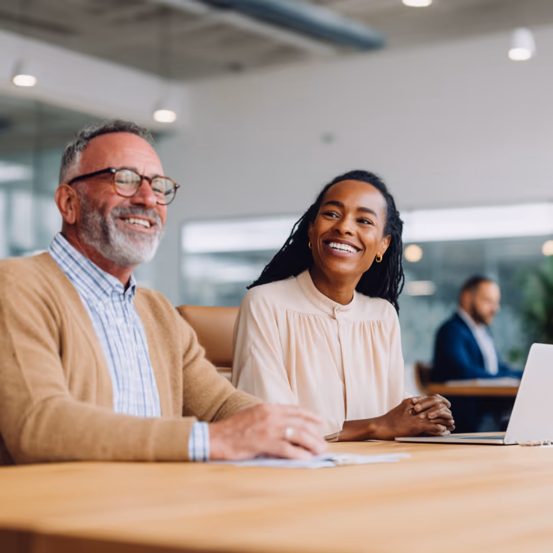 Smiling man with glasses and a woman with dreadlocks sitting and talking at a table with a laptop in a modern office.