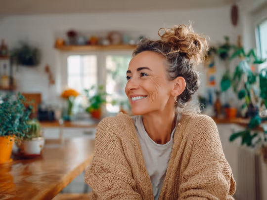 Smiling woman with curly hair in a bun wearing a beige sweater sitting at a wooden table in a bright, plant-filled kitchen.