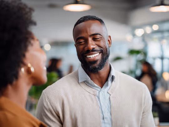 Smiling man in a light-colored sweater talking with a woman in a blurred indoor setting.