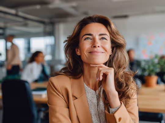 Smiling woman with long hair wearing a tan blazer sitting thoughtfully in a modern office.