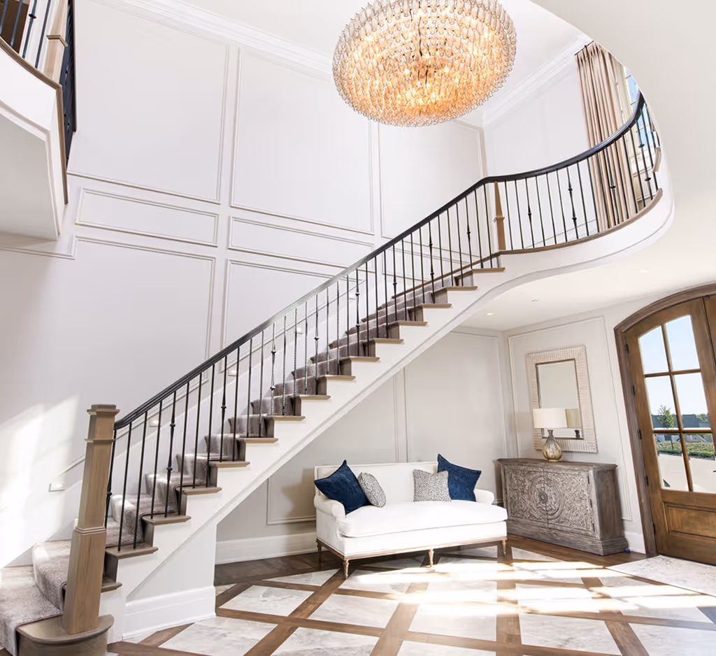 Elegant curved staircase with dark wooden railing and carpet runner above a white sofa with blue and grey cushions in a bright foyer with patterned floor and chandelier.