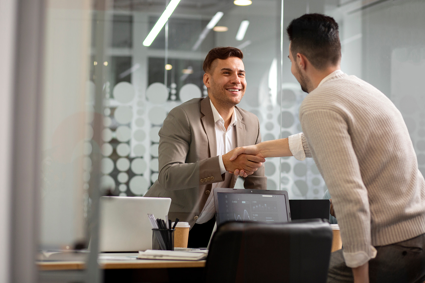 Two men shaking hands in a modern office with laptops and documents on the table.