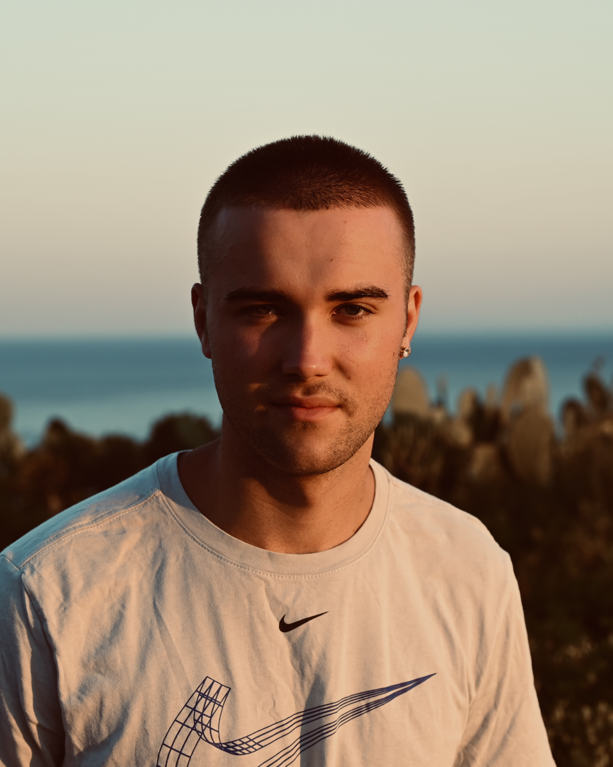 Young man with short hair and earring wearing a white Nike shirt outdoors at sunset by the sea.