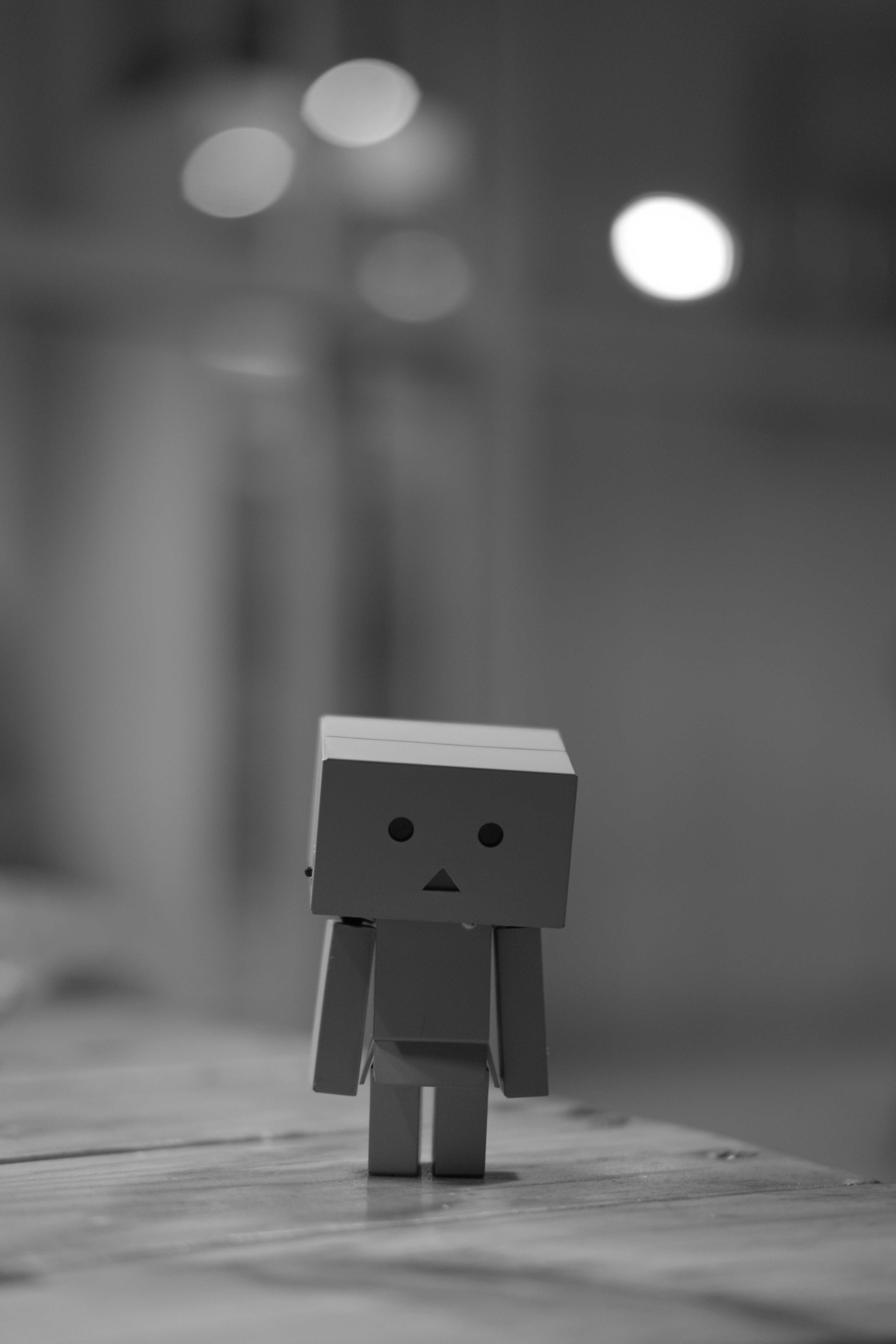 Small box-shaped robot figure standing on a wooden surface with blurred lights in the background.